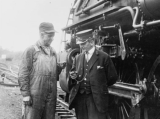 A railroad conductor and engineer looking at their pocket watches beside a locomotive circa 1930.