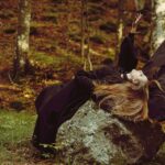 Photo of a woman dressed as a witch laying on a rock in a forest.