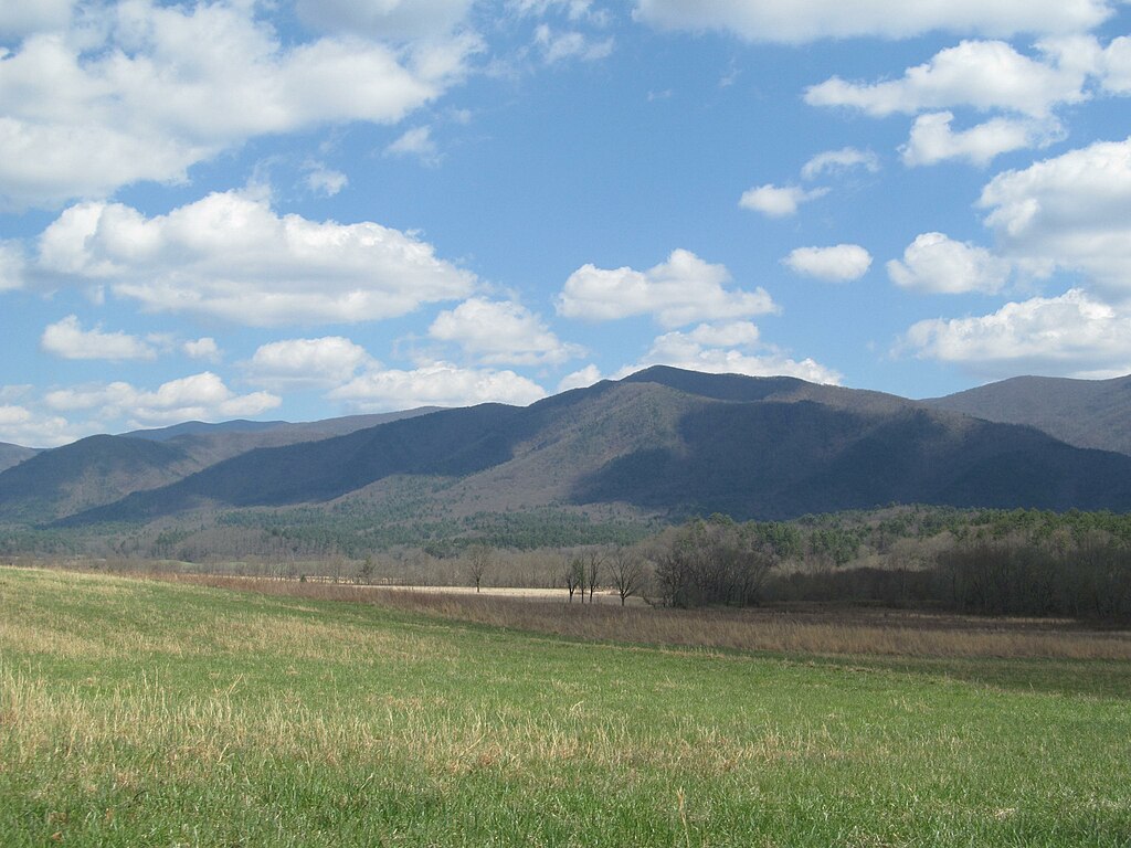 View of a meadow surrounded by mountains with clouds casting shadows.